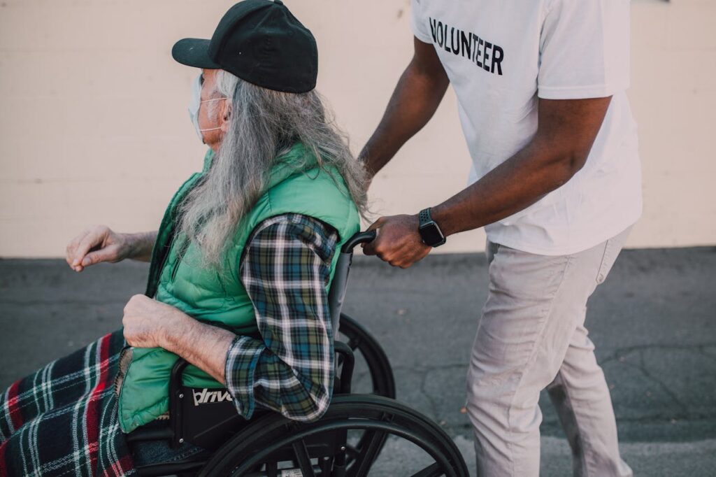 pexels-photo-6646975 A volunteer helps a senior in a wheelchair during daytime outdoors.